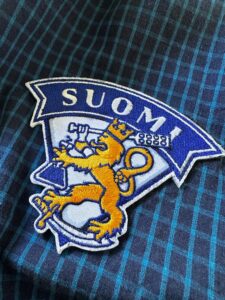 Brodeerattu kangasmerkki, jossa Suomen teksti "Suomi" sekä leijona, jolla on kädessään mallasmela ja erlenmeyerpullo