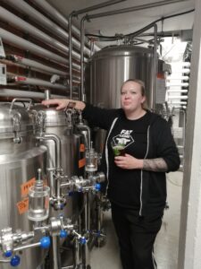 Head brewer Greta Mäkinen käymisastian vieressä, kädessään lasillinen olutta